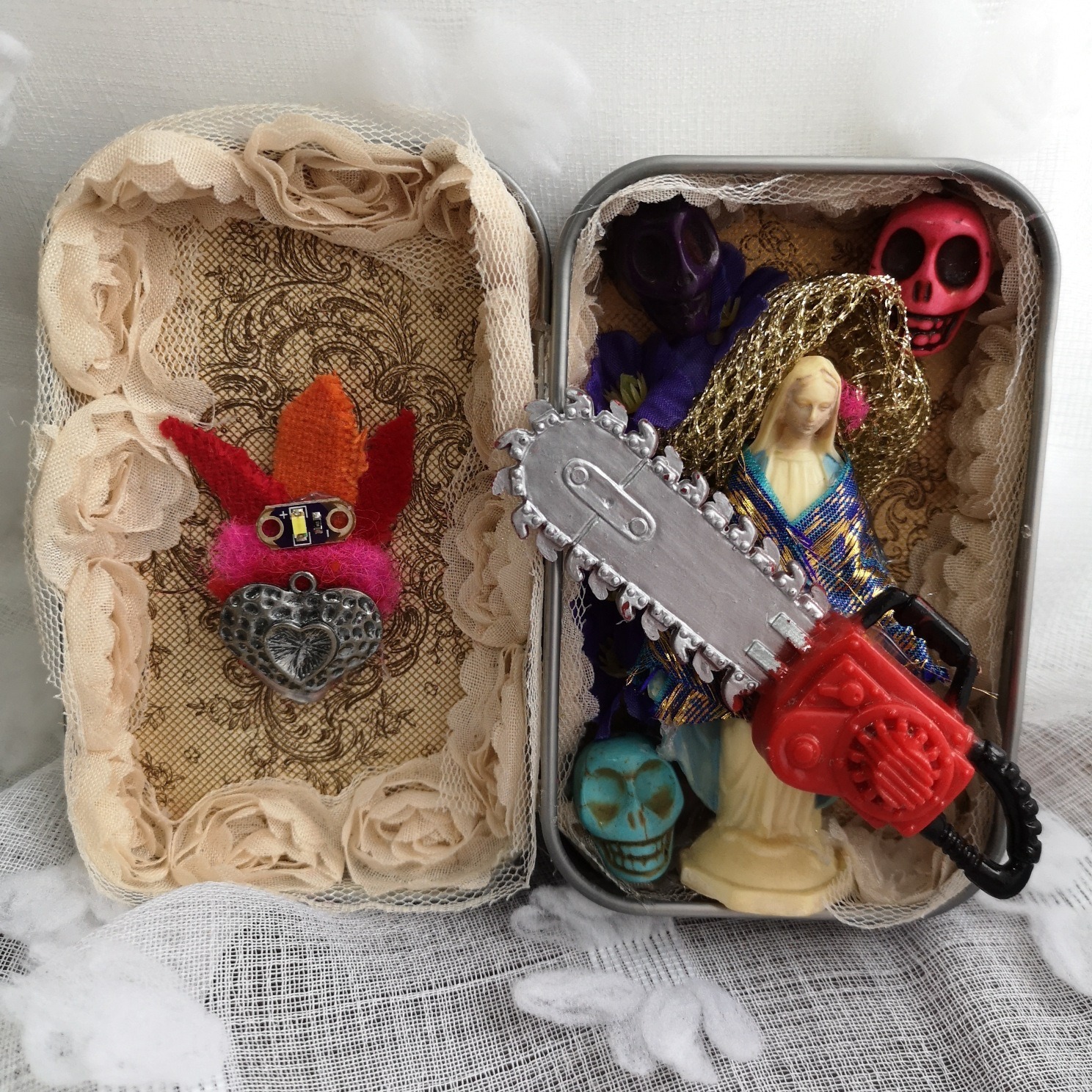 open rectangular metal tin decorated with paper, ribbon, a miniature statue of Mary holding a chainsaw and surrounded by skull beads